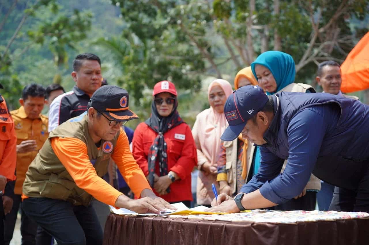 Patahudding membungkuk menandatangani dokumen di meja dengan disaksikan petugas dan peserta apel