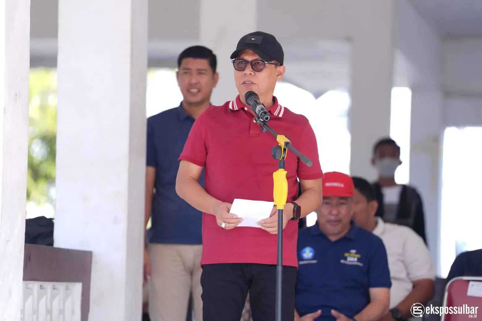 Junda Maulana memakai kaus merah dan topi hitam sambil berbicara di depan mikrofon